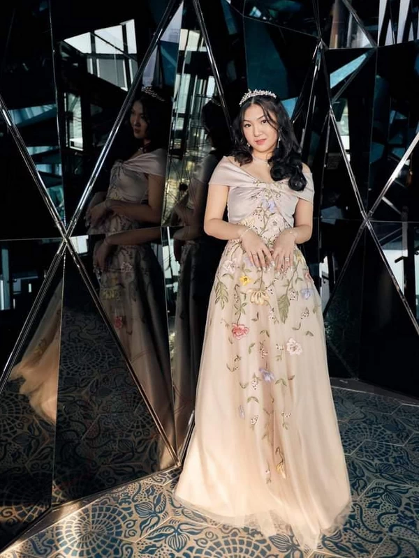 Wanita muda mengenakan gaun panjang warna peach dengan motif bunga-bunga timbul warna-warni. Gaun dibuat oleh Fifilia.